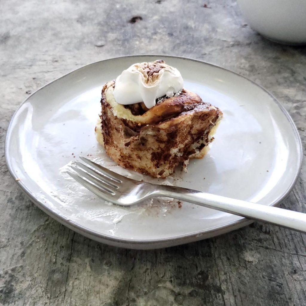 smores cinnamon rolls with marshmallow meringue side view one bun on a plate with fork for a view of swirls inside light gray plate on distressed gray wood countertop meringue is toasted