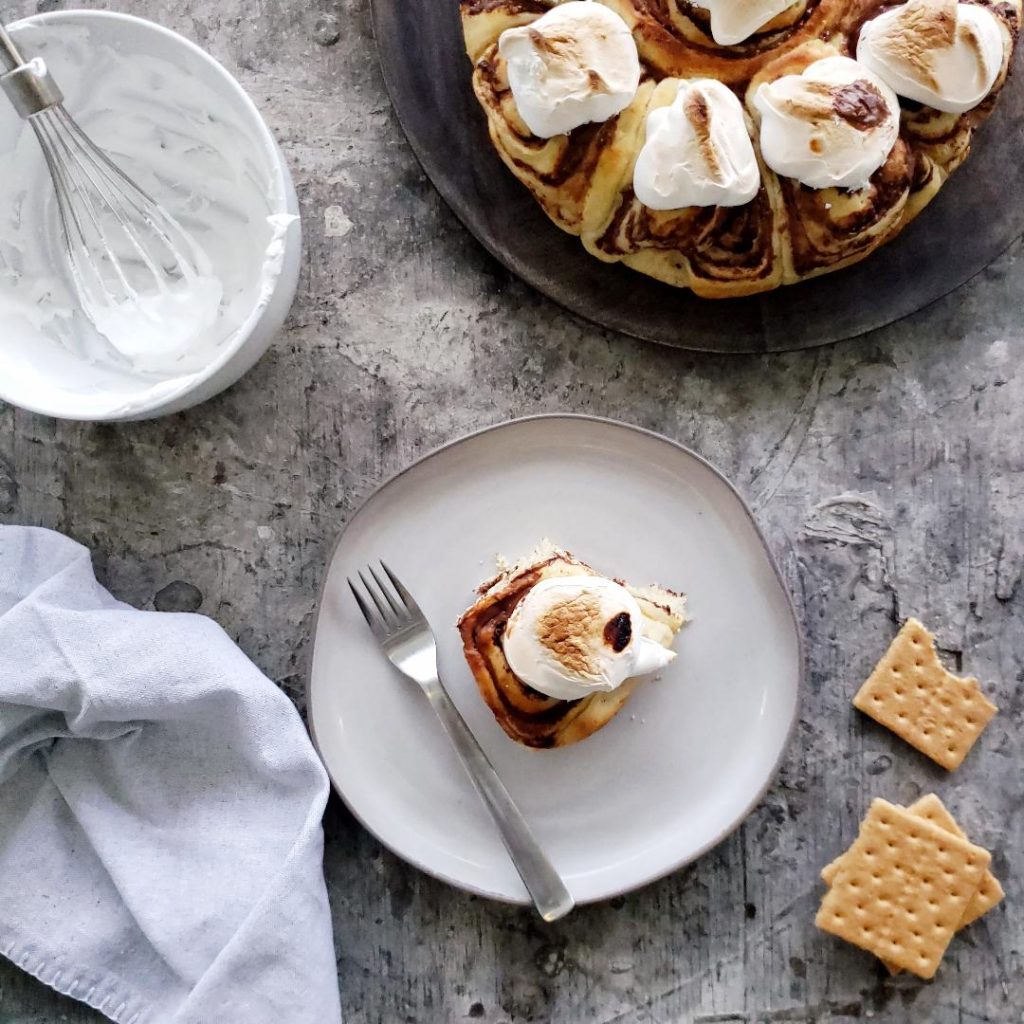 s'mores cinnamon rolls with marshmallow meringue top down photo distressed gray table light gray plate with one smores roll with toasted marshmallow meringue on top and a single fork top right of photo is a round black charger plate with the remaining batch of smores rolls top left is a white bowl with a metal whisk and marshmallow meringue bottom left is a gray linen and bottom right is three graham crackers