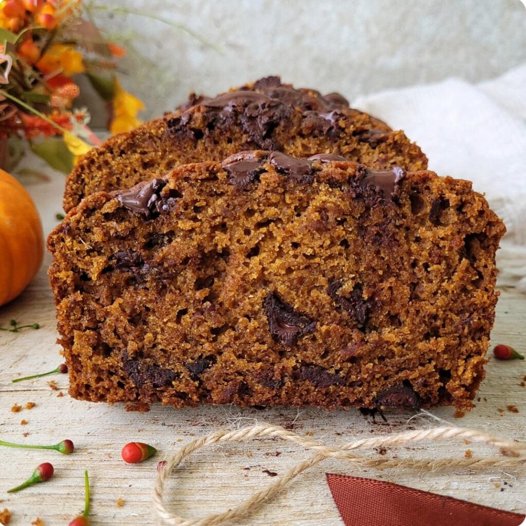 pumpkin chocolate chip bread. side view of a slice so you can see the inner crumb and chocolate chips. background has a small pumpkin and fall colored flowers. 