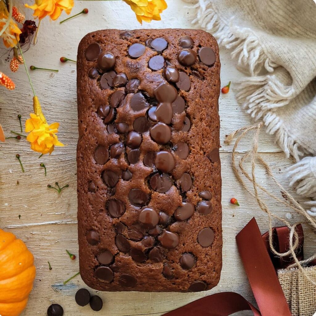 pumpkin chocolate chip bread. top down view of uncut loaf. background is styled with fall flowers, twine and cream colored fringe. 